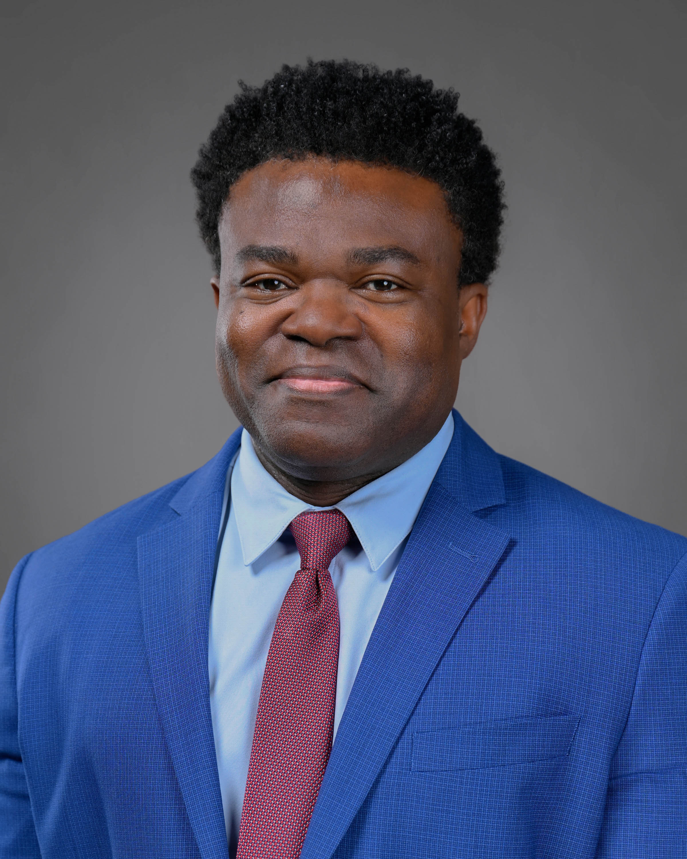 Kind man smiling with curly dark hair wearing a blue jacket with a light blue shirt and red tie.